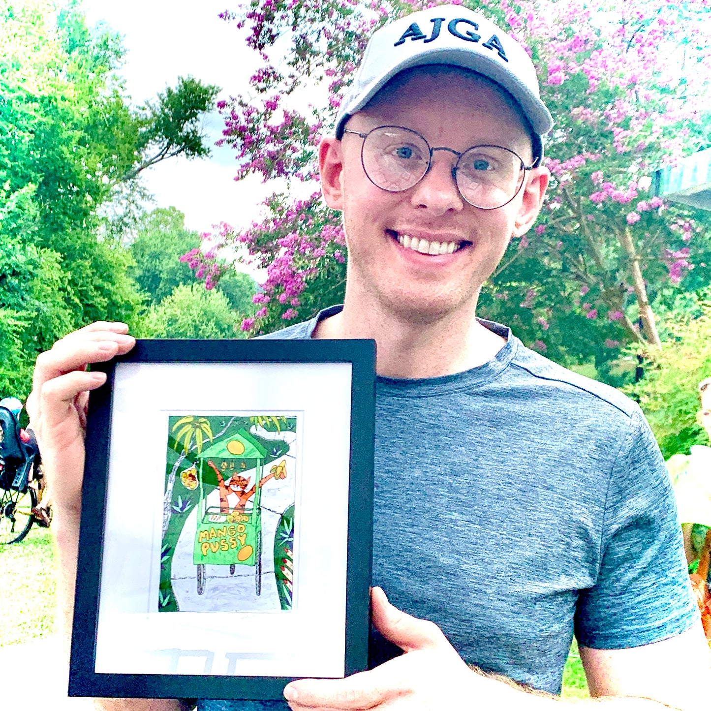 a man holding a framed art print featuring Superpussy-an orange cat-pushing a mango cart down a sidewalk in the jungle. The cart says "Mango Pussy" on the front.