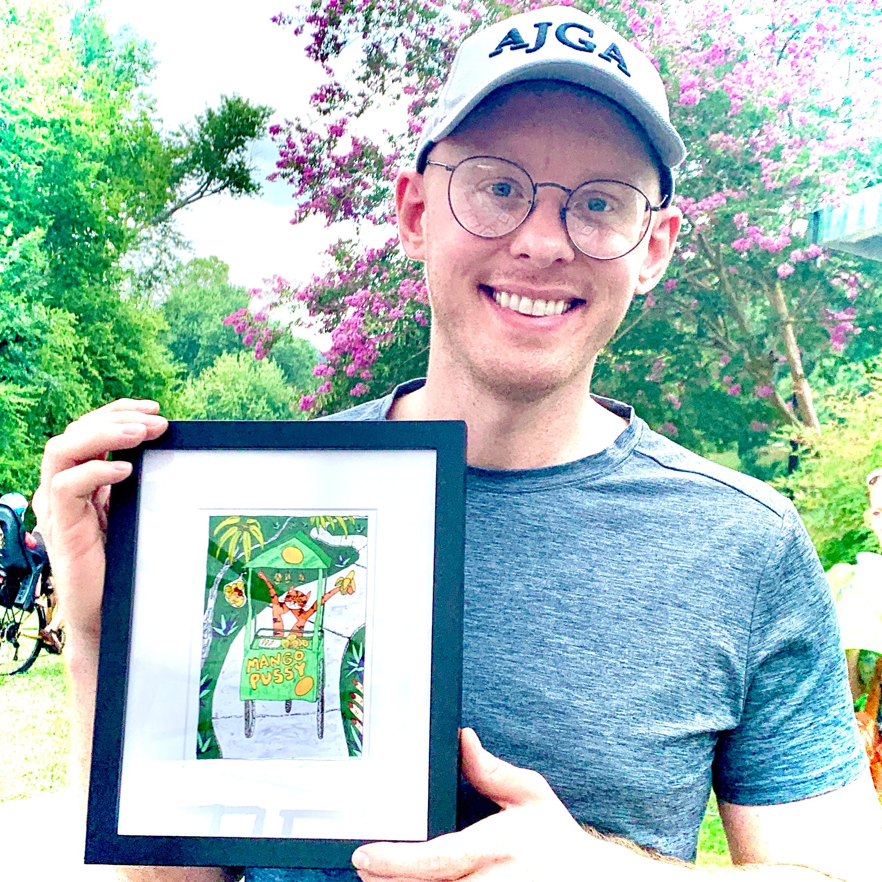 a man holding a framed art print featuring Superpussy-an orange cat-pushing a mango cart down a sidewalk in the jungle. The cart says "Mango Pussy" on the front.